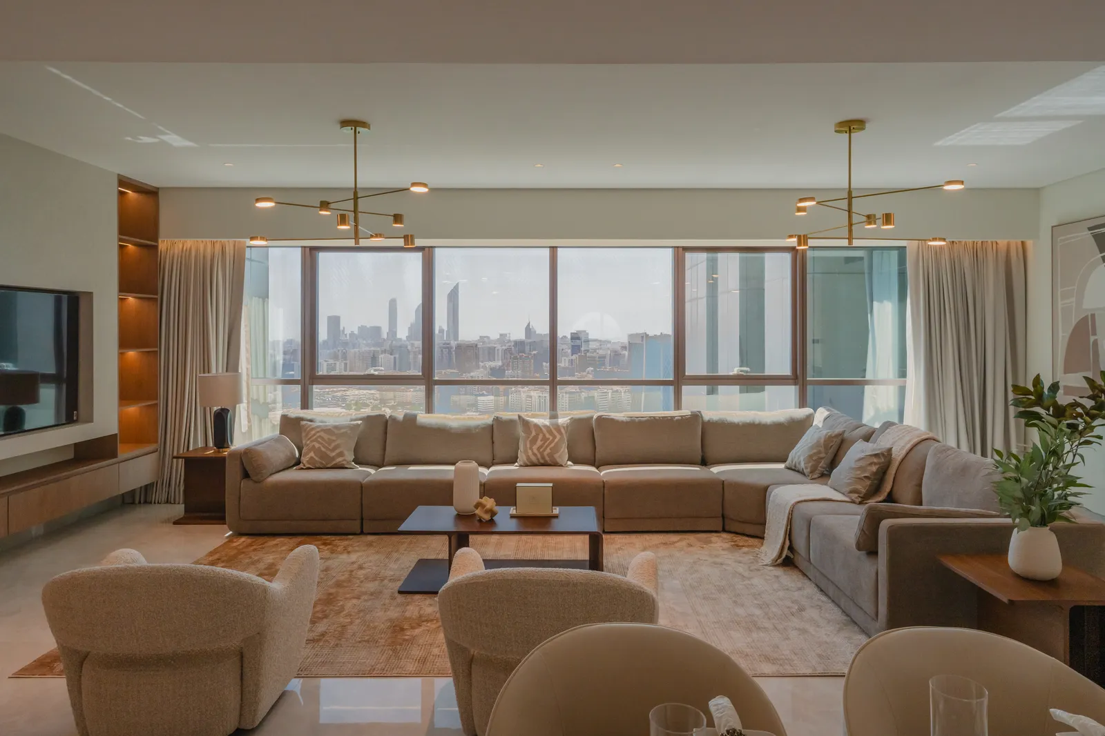A furnished living room: a tan sectional sofa facing floor-to-ceiling windows onto the Abu Dhabi skyline across the water, two brass branched ceiling lights, boucle armchairs, a television wall with lit shelving and a polished marble floor