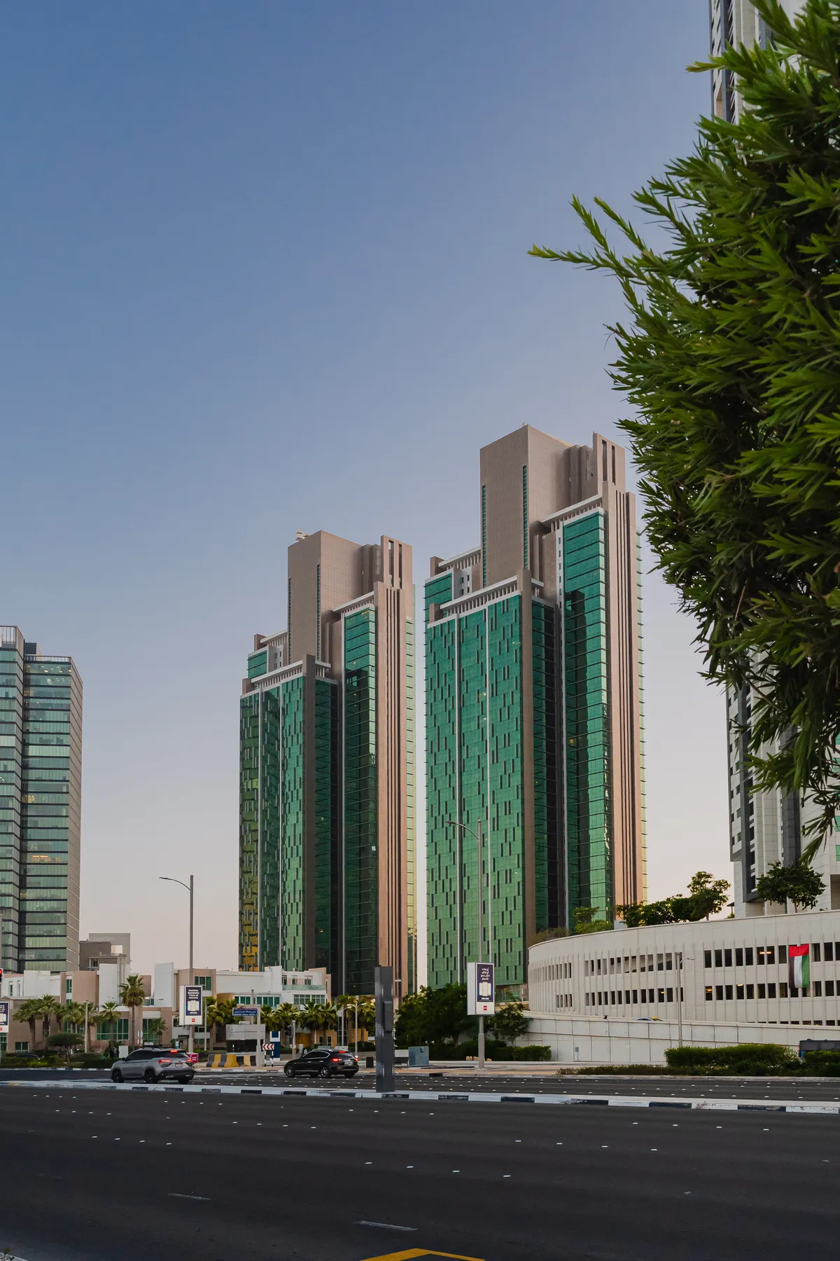 Al Durrah Tower and the matching MAG 5 Residence tower seen across a road in Marina Square, their stepped stone crowns against a clear evening sky, a tree in the foreground and a cream building with a UAE flag at their base