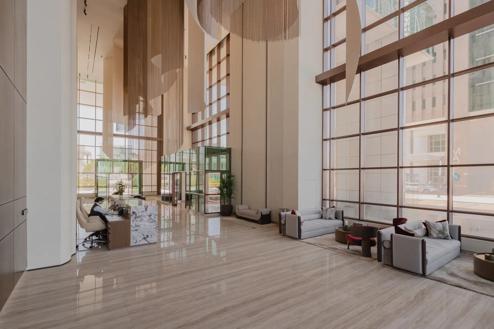 The lobby looking towards the entrance: a receptionist at a marble desk, grey sofas with burgundy cushions on a rug beside the tall window wall, chain curtains hanging above