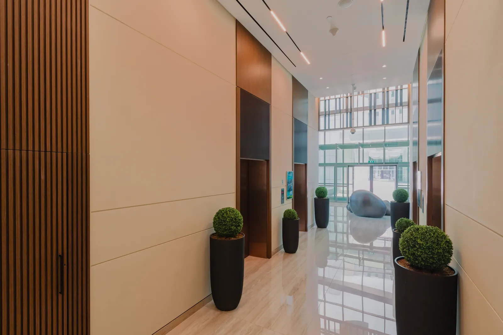 The lift lobby from the side: two bronze lift doors, box plants in black planters, the corridor opening onto the double-height glazed lobby