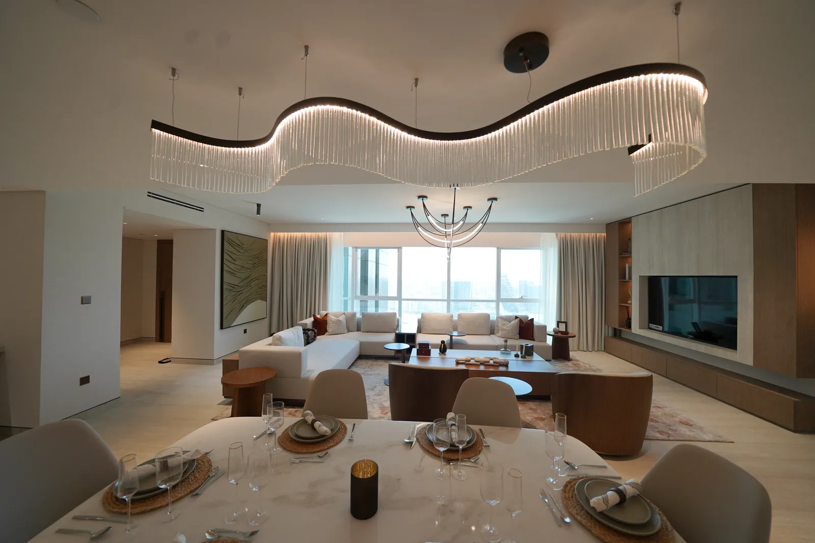 The dining table laid under a wave-shaped crystal chandelier, with the living room's branched pendant, sofas and window wall beyond
