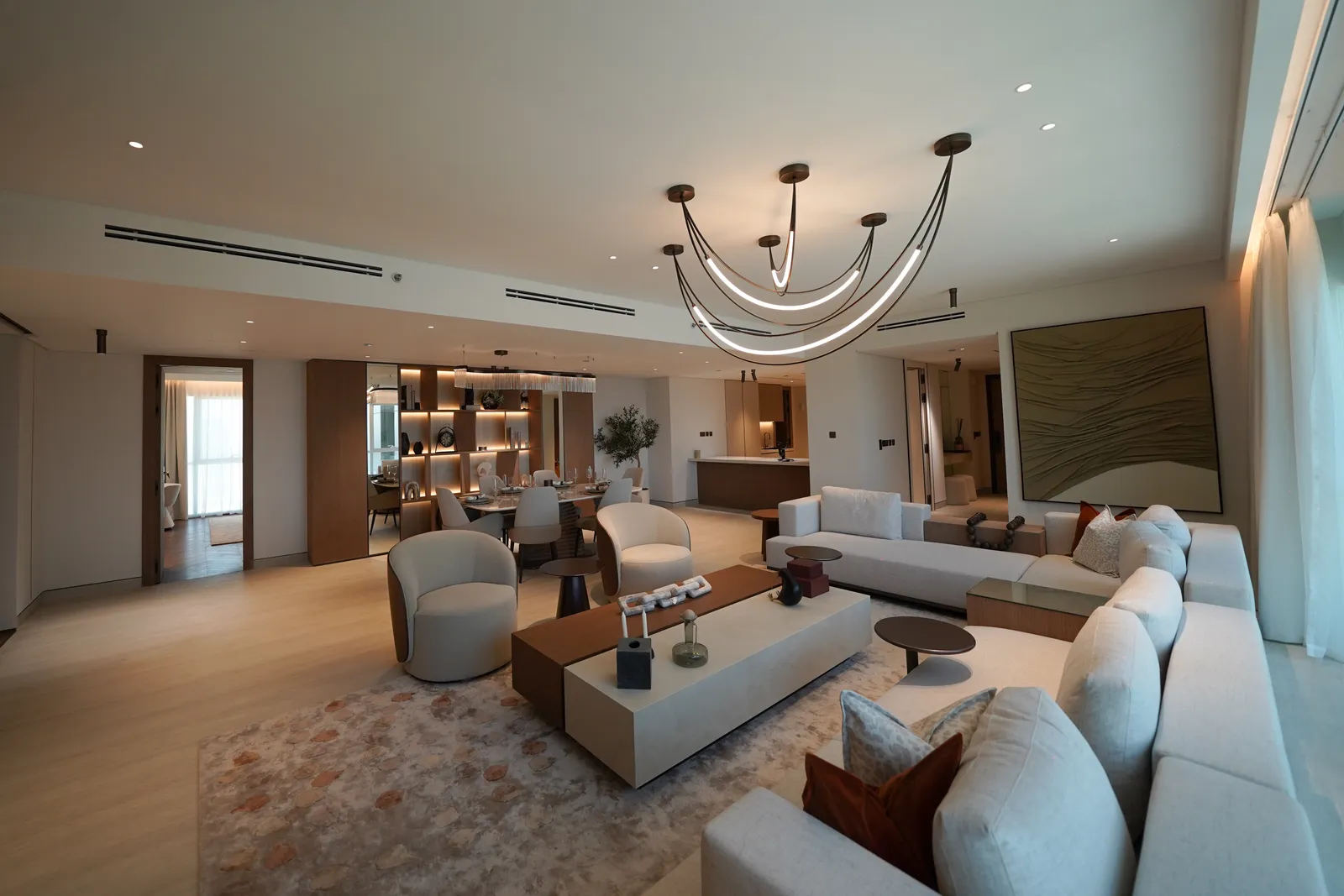 The living room: cream sofas and tub chairs around a two-tone coffee table on a rug, a branched chandelier, an abstract painting, the dining area and kitchen counter beyond