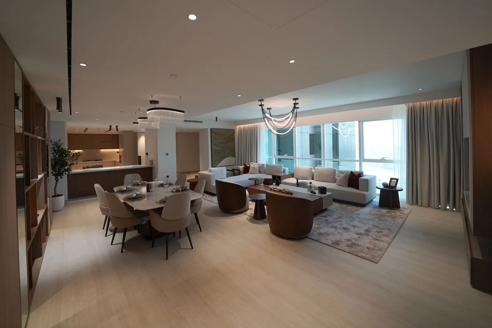 The open-plan room from the shelving side: dining table, the branched pendant, sofas and the window wall, the kitchen with an olive tree at left