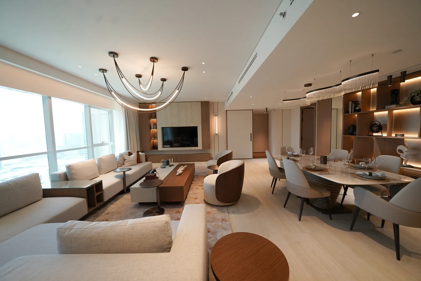 The living room with sheer curtains across the window wall, the branched chandelier, the television wall, sofa and dining table under crystal pendants