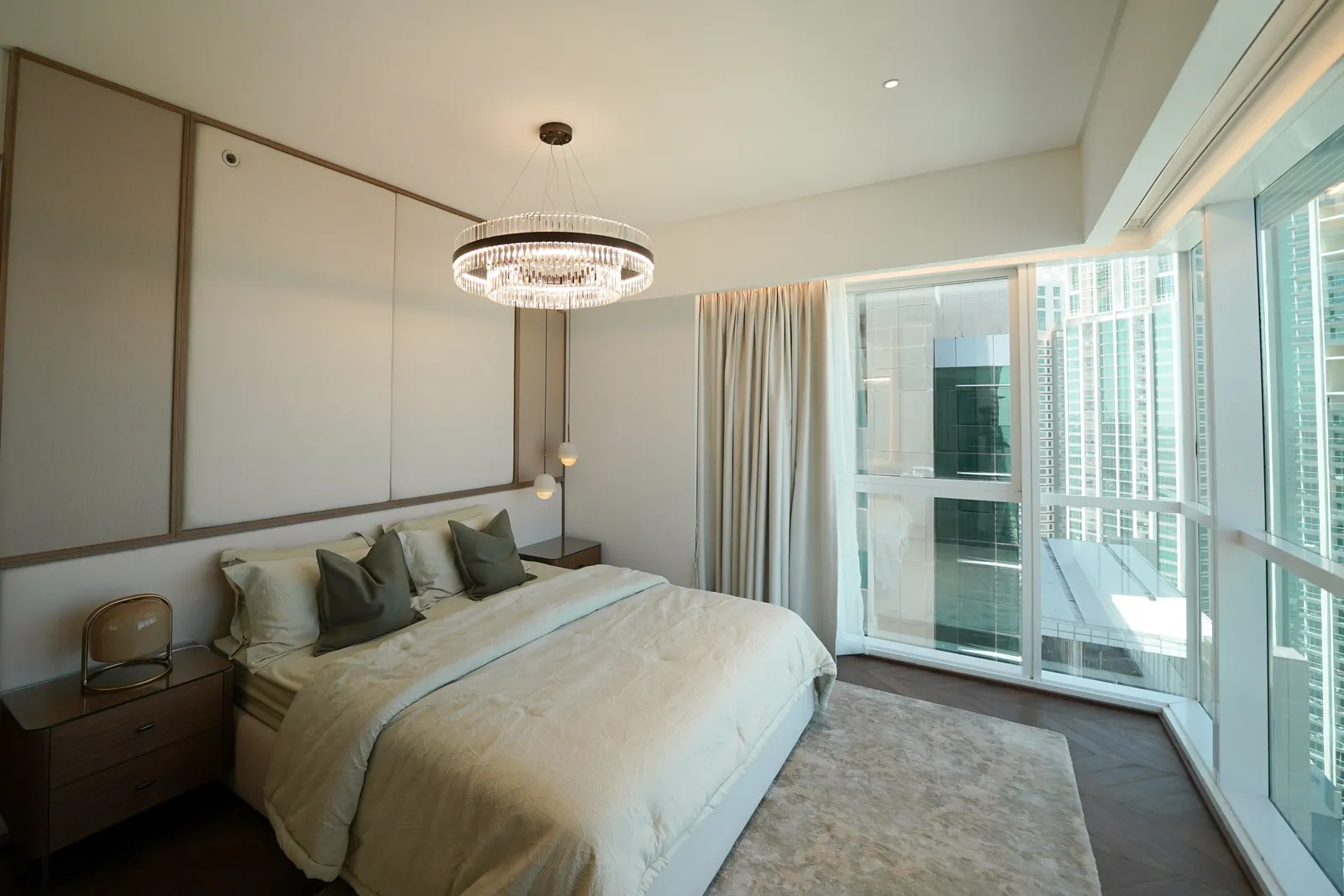A bedroom with a crystal ring chandelier and a double bed with green cushions beside a corner window onto neighbouring towers