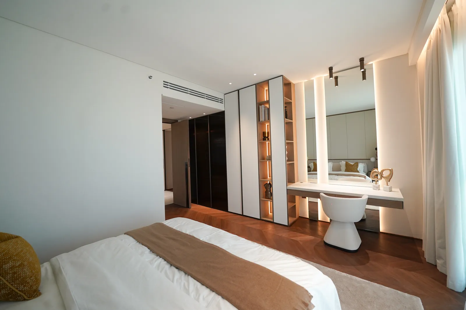 A dressing area off a bedroom: a lit mirror, shelving and a white stool, dark wardrobes and a herringbone floor