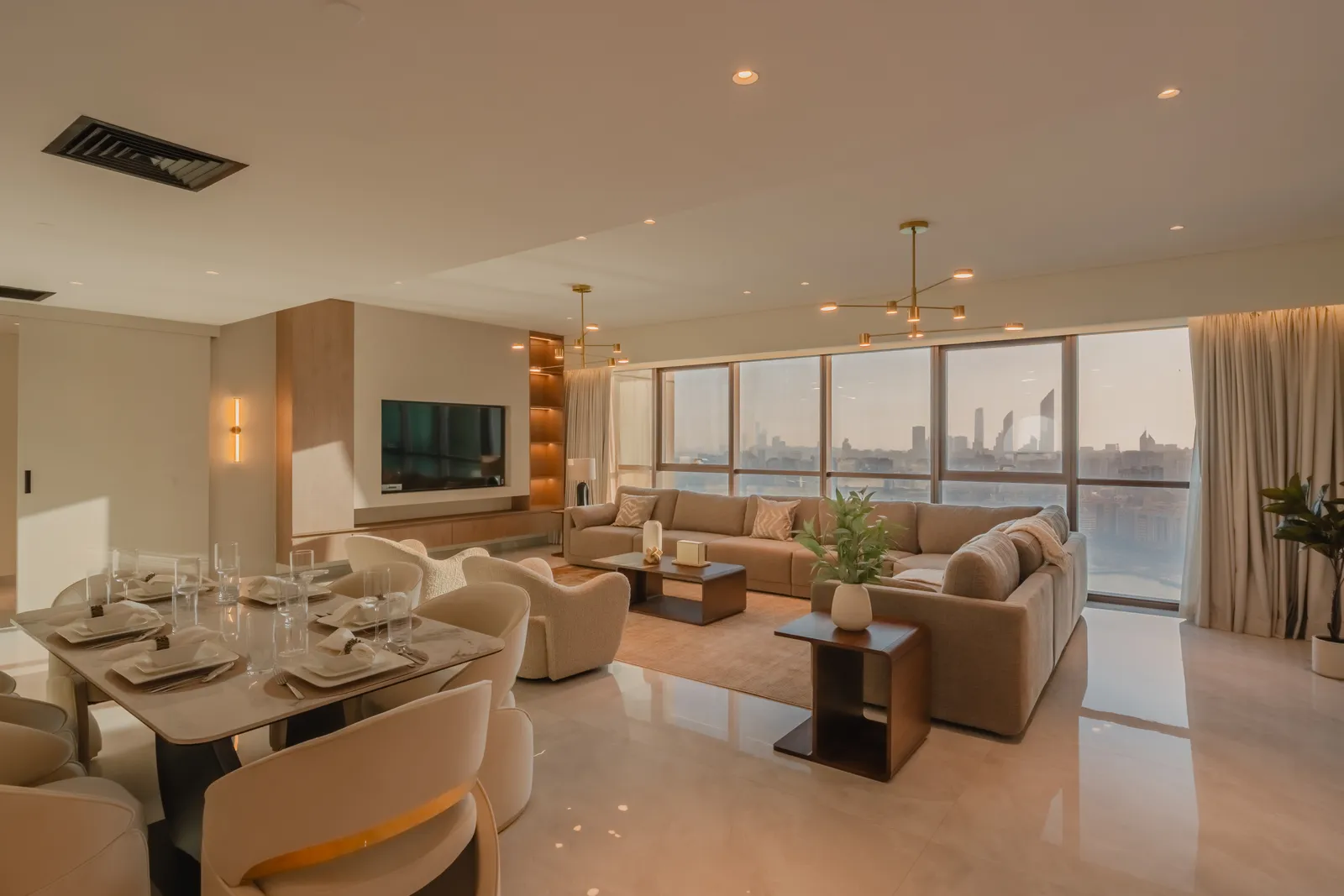A living and dining room in evening light: a dining table laid for six with boucle chairs, the tan sofa, brass ceiling lights and the skyline through the windows
