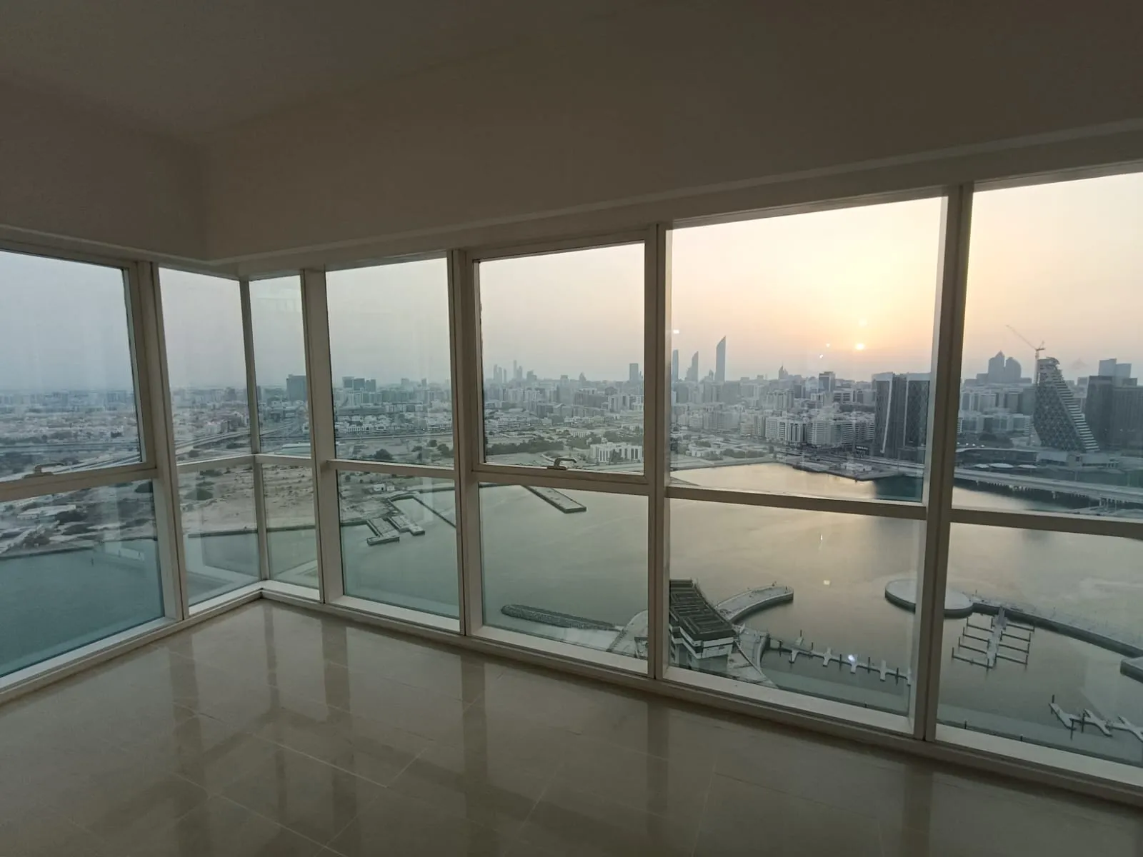 An unfurnished apartment with floor-to-ceiling glazing on two sides, looking over the marina and the water to the Abu Dhabi skyline at sunset