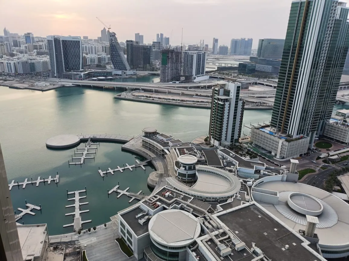 The marina from a high floor at sunset: pontoons and moored boats, round-roofed buildings at the water's edge, a bridge and the city skyline