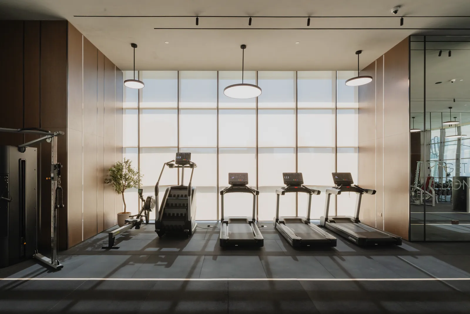 The gym: three treadmills and a stair climber in front of a floor-to-ceiling window with blinds, round pendant lights, a cable machine at left and a mirrored wall at right
