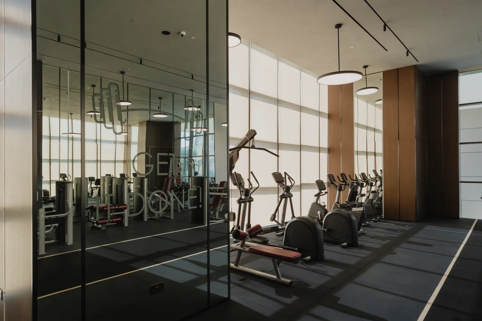 The gym: a mirrored wall lettered GET IT DONE!, cross-trainers, bikes and a bench along the window wall, a timber-clad column