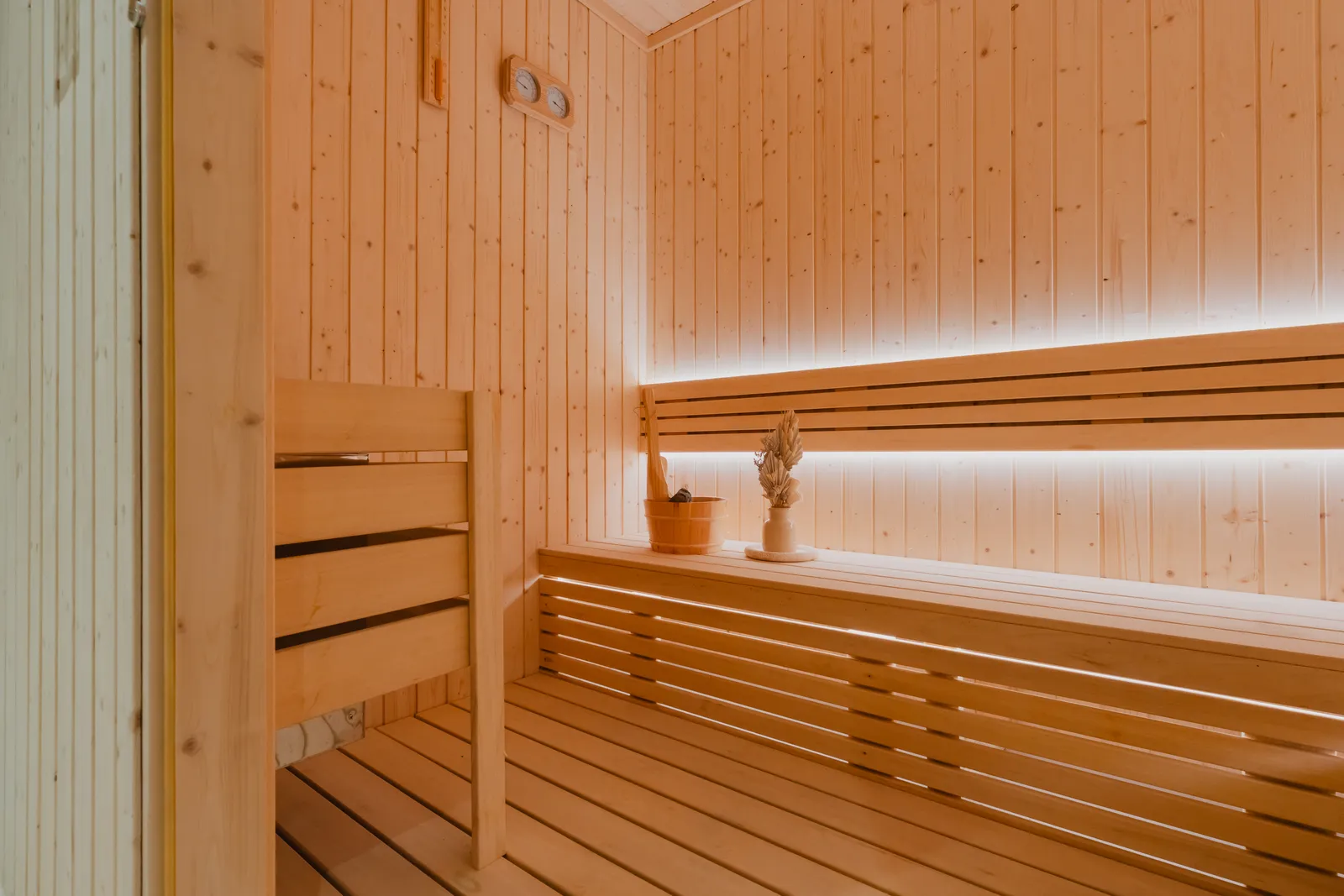 The sauna from its door: pale timber walls and tiered benches, a bucket and ladle on the upper bench, a thermometer and hygrometer on the wall, light under the benches