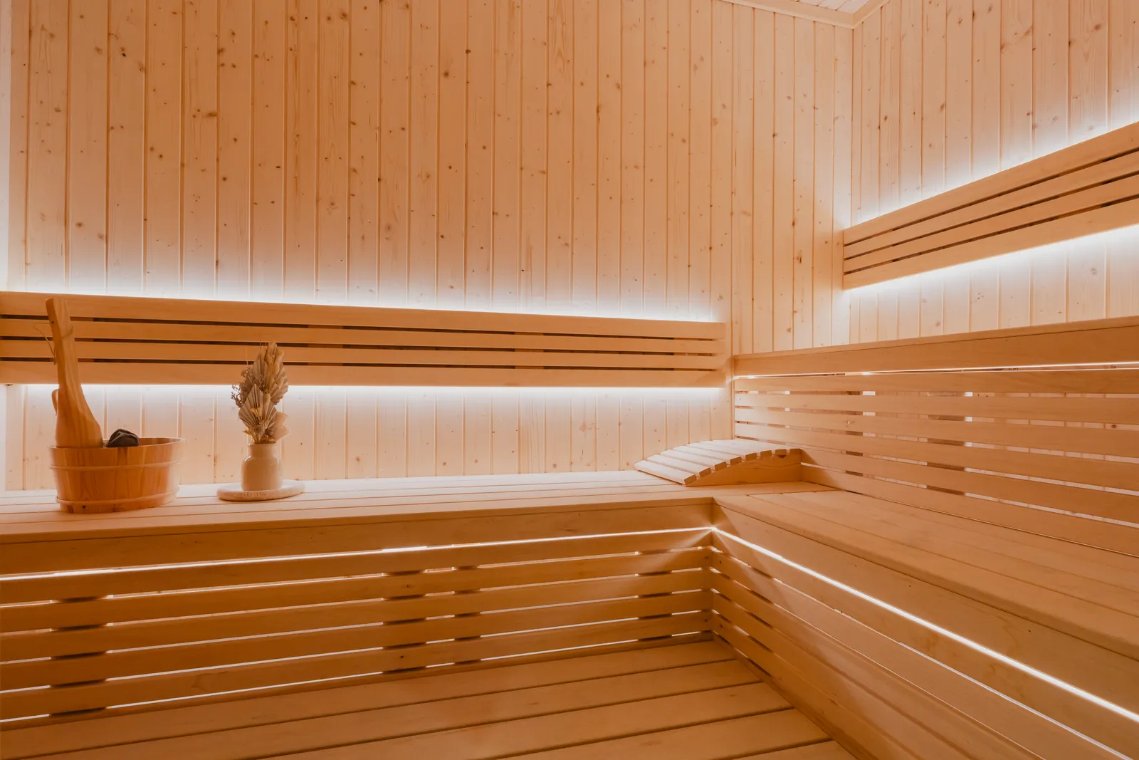 The sauna: tiered pale timber benches with a bucket, a ladle and a vase of dried grass, lit from behind the slats