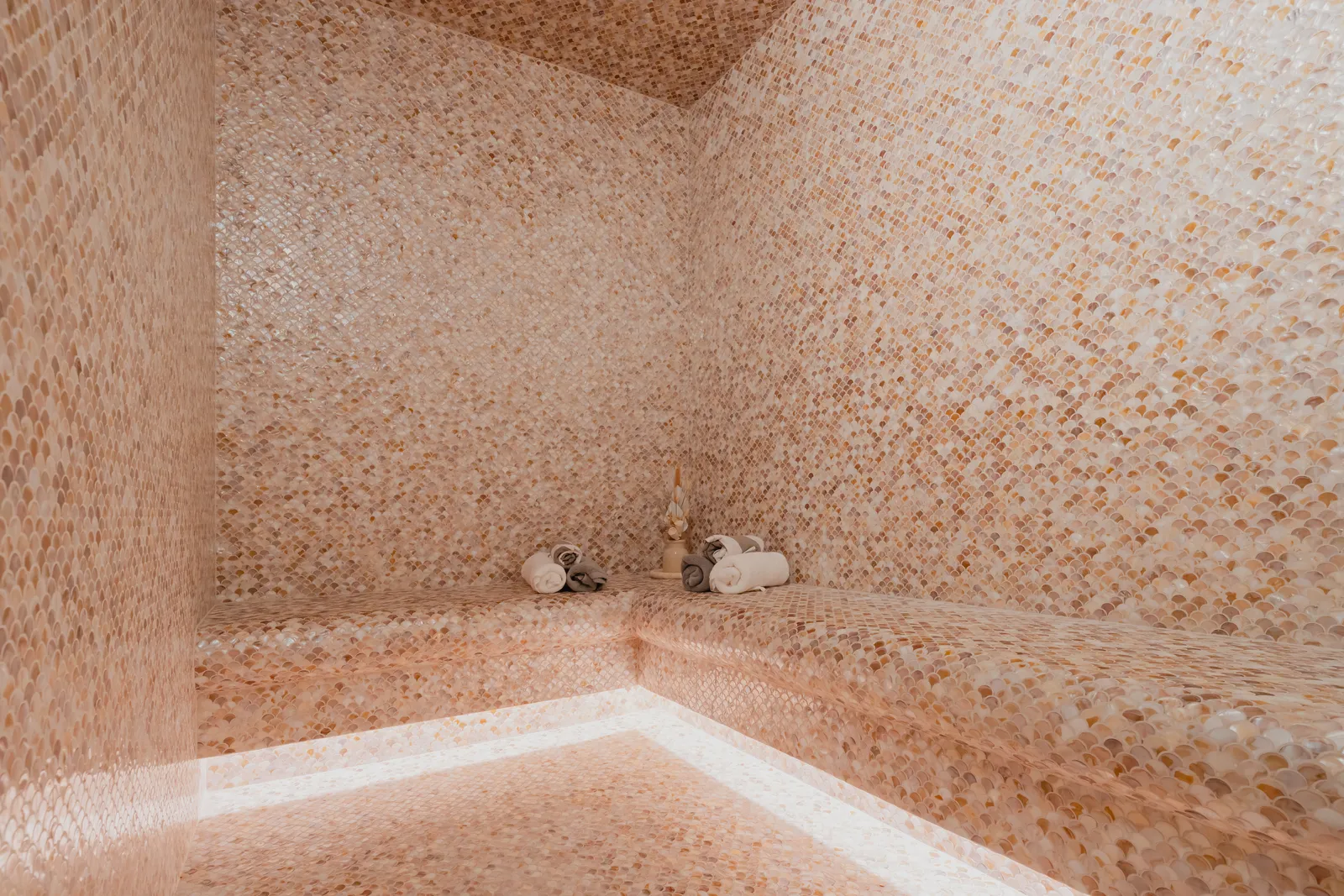 The steam room: mosaic-tiled walls and benches in pink and cream with rolled towels in the corner, lit from under the bench