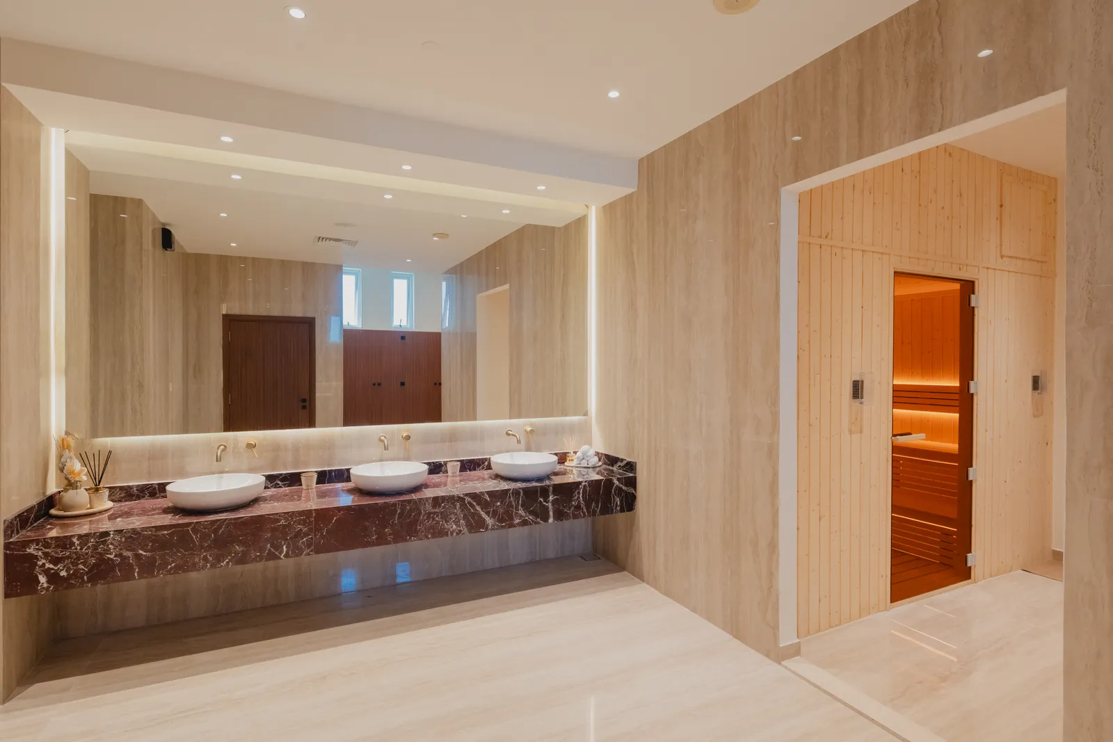 The vanity's red marble counter and basins under the backlit mirror, with the sauna's timber entrance and glass door at right