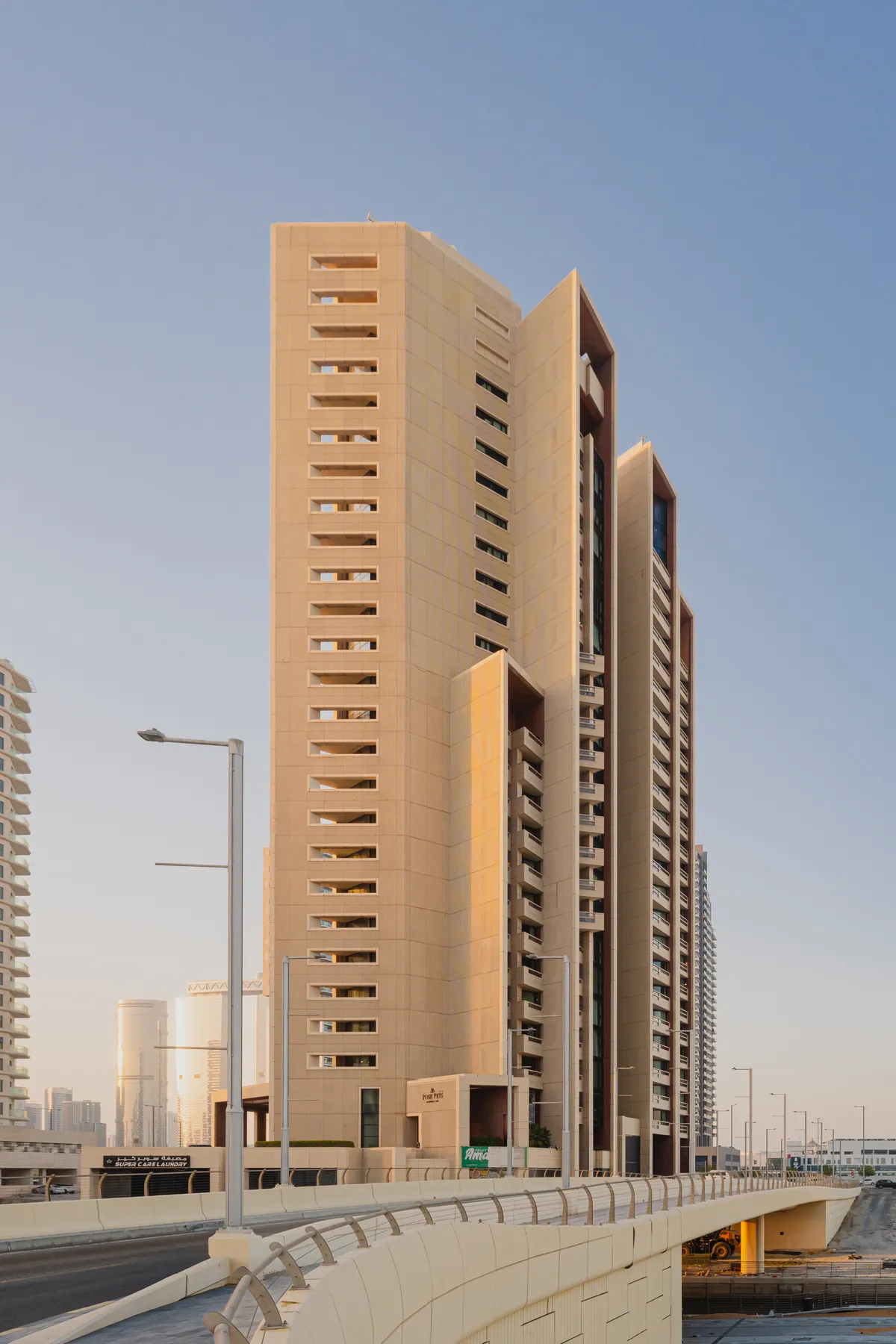 Baheen Tower from the road below in evening light: the full height of the sand-coloured tower, its stepped profile and recessed balconies, with other Al Reem Island towers behind