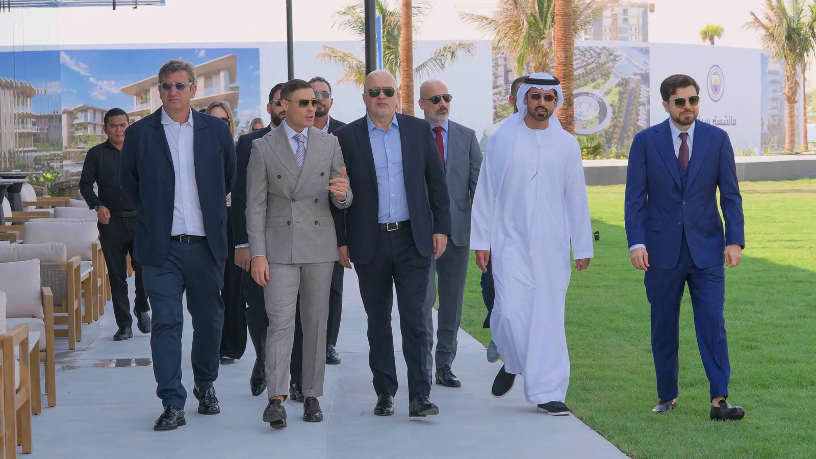 A group of men in suits and kandura walking along a paved path beside the canal, hoardings printed with renders of the homes behind them