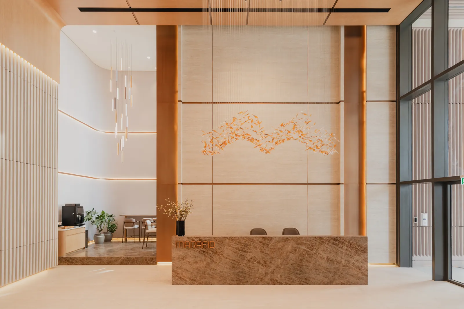 The reception lobby: a brown marble desk lettered MARAFID in bronze, under a travertine wall with a swirl of gold leaves, a cascade of pendant lights and the coffee corner to the left