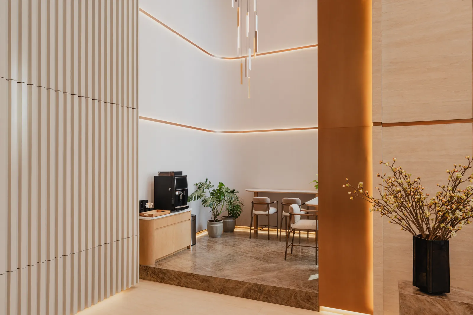The coffee corner off the lobby: an espresso machine on a timber counter, two small tables with chairs, a bronze column, a tall vase of branches and pendant lights