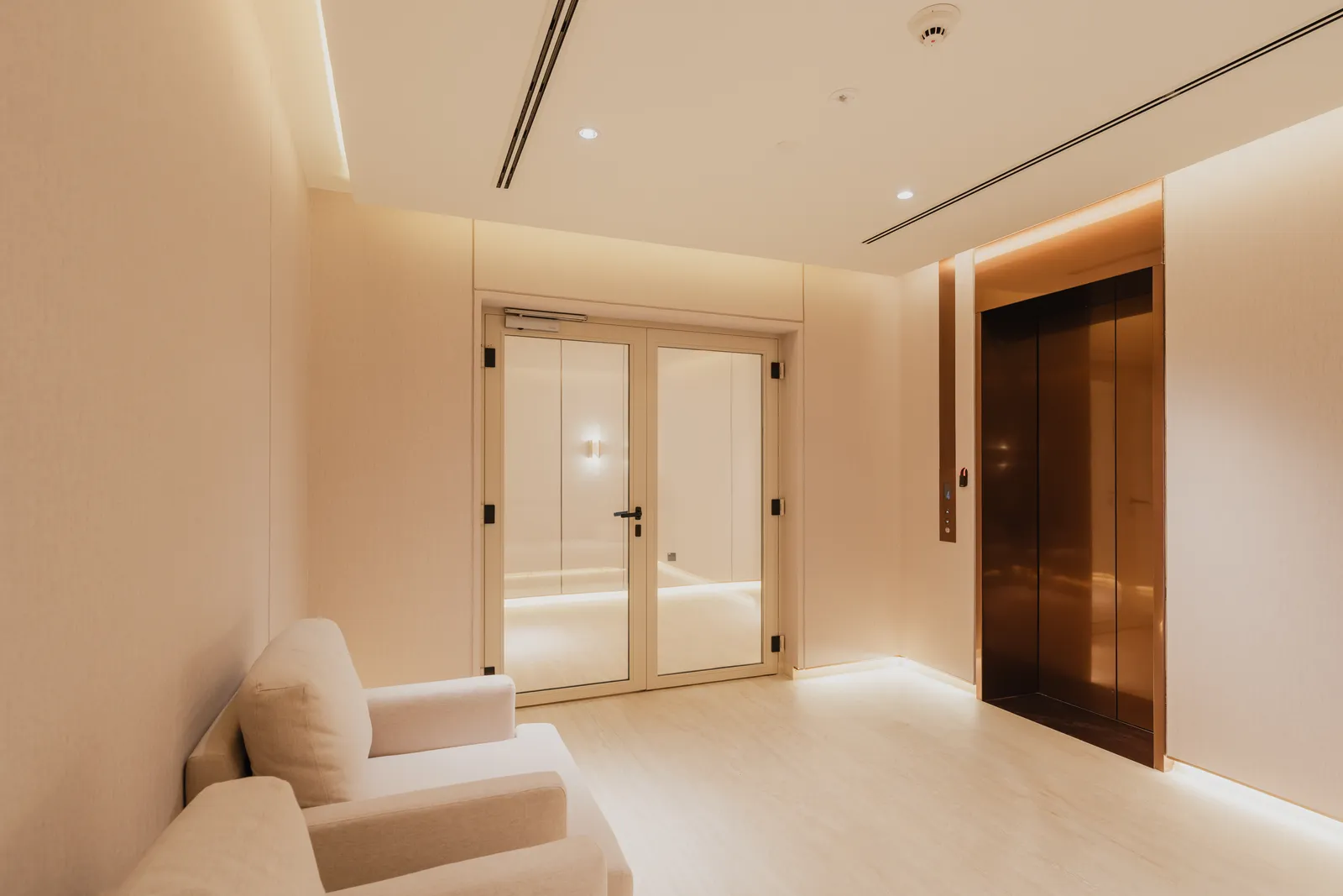 The private lobby of a three-bedroom apartment: glazed double doors into the home, the bronze lift beside them and two cream armchairs